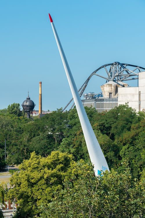 Zwischen grünen Bäumen ragt ein riesiges, weißes Rotorblatt senkrecht hervor. Im Hintergrund sind die gebogenen Träger eines Gebäudes, ein Wasserturm und ein Schornstein erkennbar.