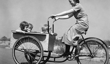A woman rides a cargo bike. Four toddlers are sitting in the large open box in the front of this three-wheeled bike. The black-and-white photograph shows this scene from the side, with the woman biking from right to left.