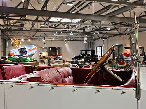 Einblick in den offenen Innenraum eines historischen Automobils. Das Fahrzeug ist weiß lackiert, die Sitze sind mit rötlichen Leder bezogen und das Lenkrad aus Holz. Im Hintergrund befinden sich weitere Fahrzeuge. Ein orange-blauer Rennwagen ist an einer Wand befestigt.