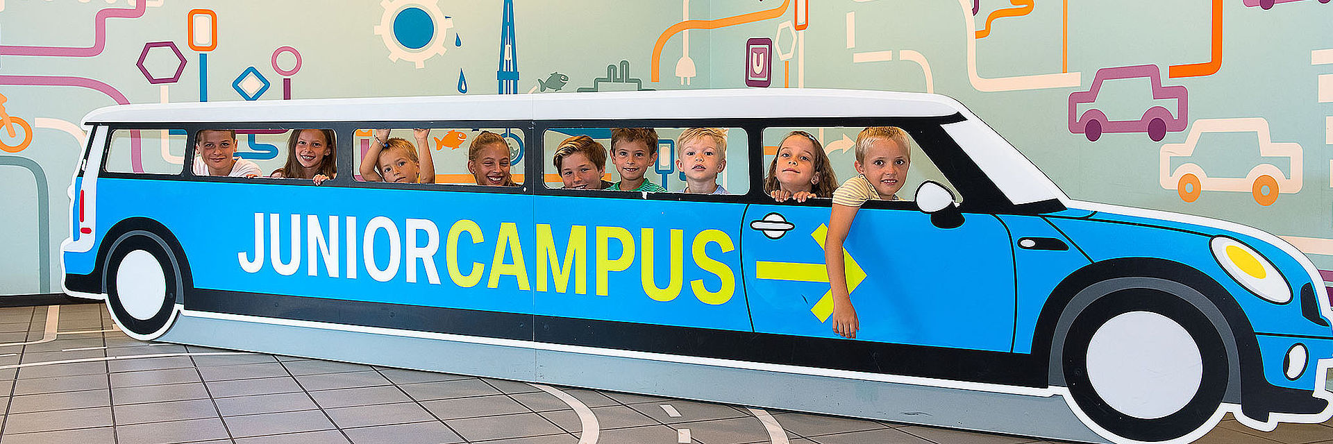 Neun Kinder schauen durch die Seitenfenster eines etwa sechs Meter langen Aufstellers aus Pappe in Form eines Autos. Auf der Seite ist der Schriftzug „Junior Campus“ in grün auf blauem Grund angebracht. Auf dem Boden sind Fahrbahnmarkierungen angedeutet.
