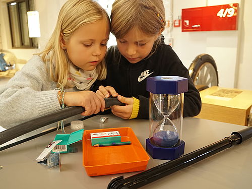 Zwei Kinder flicken in der Ausstellung einen Fahrradschlauch. Auf dem Tisch befinden sich eine Luftpumpe, eine Reifenreparatur-Set und eine Sanduhr.