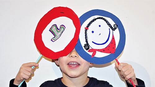 Ein Junge hält sich zwei selbstgemalte Verkehrschilder vor die Augen. Das eine Schild ist rot umrandet, das andere blau umrandet und zeigt ein fröhliches Gesicht.
