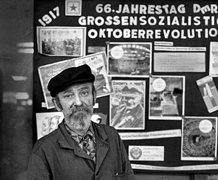 Auf dem Foto ist ein Arbeiter mit Lenin-Mütze zu sehen, der vor einem schwarzen Brett mit kommunistischen Parolen und einem Foto von Lenin steht.