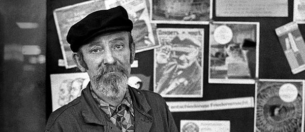 Auf dem Foto ist ein Arbeiter mit Lenin-Mütze zu sehen, der vor einem schwarzen Brett mit kommunistischen Parolen und einem Foto von Lenin steht.