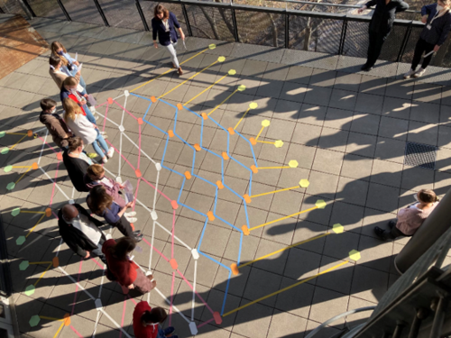 Eine Grupper erwachsener Personen steht auf einer Terrasse. Auf dem Boden ist ein Netz aus verschiedenen Farben aufgemalt.