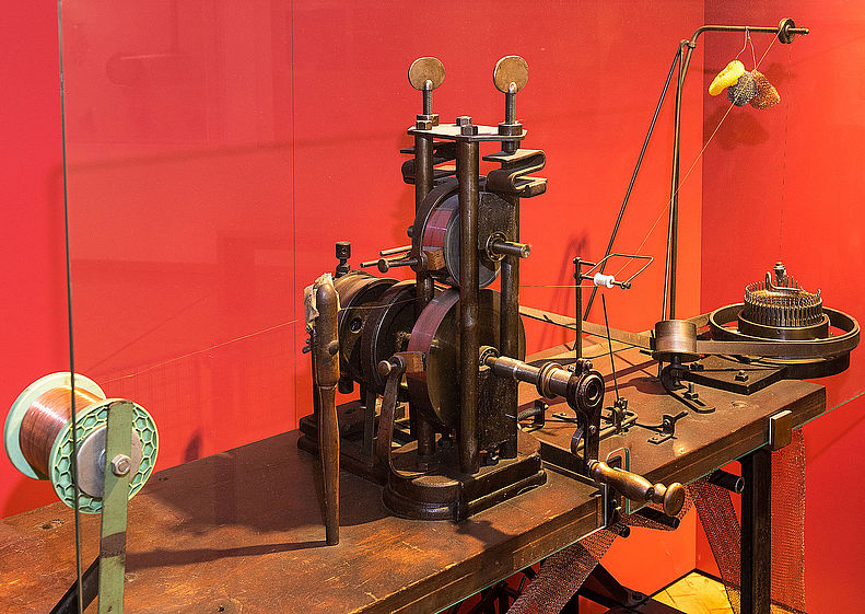 Die Strickmaschine aus Metall steht in der Mitte einer hölzernen Werkbank. An der Vorderseite sitzt eine Handkurbel. Links der Maschine steht eine weiße Spindel. Ein Metall-Faden wird durch die beiden übereinanderliegenden Räder der Maschine geführt.