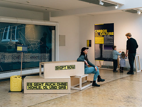Mehrere Menschen halten sich in einem Ausstellungsbereich auf. Auf Sitzmöbeln steht "Shift now" und "Es geht auch ohne Kohle, Öl und Gas". Auf einem Plakat steht "Carbon Culture". Außerdem ist ein gelber Kanister zu sehen.