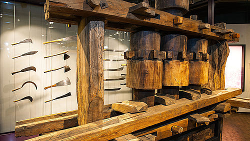 Eine große, hölzerne Zuckerrohrmühle. Sie besteht aus einer langen Antriebsstange oben, die eine Antriebswalze zum Rotieren bringt. Diese setzt zwei weitere Walzen in Gang, die das Zuckerrohr auspressen.