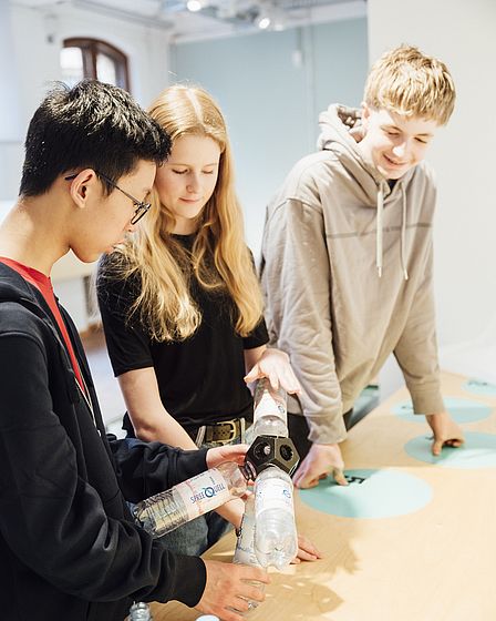 Ein Mädchen und ein Junge schrauben gemeinsam leere Plastikflaschen in eine Kunststoff-Fassung. Das Ergebnis ist eine lustig geformte Lampe. Ein zweiter Junge steht fröhlich daneben und schaut zu.