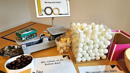 Auf einen Tisch stehen verschiedene Objekte und Zettel mit Fragen zum Thema Messen.