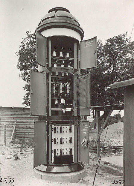 Die kegelförmige Litfaßsäule ist auf einer Seite geöffnet. Auf drei Ebenen befinden sich Türen, hinter denen die Technik des Transformators zugänglich ist. Es ist die Aufschrift „Hochspannung, Vorsicht! Lebensgefahr“ zu lesen.