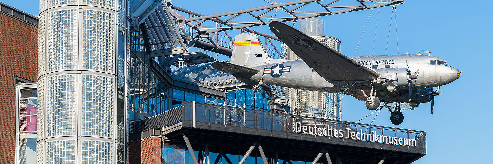 Ein modernes Neubaugebäude mit Glasfassade. An einer Stahlkonstruktion auf dem Dach hängt ein Flugzeug. 