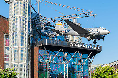 Ein modernes Neubaugebäude mit Glasfassade. An einer Stahlkonstruktion auf dem Dach hängt ein Flugzeug. 