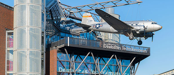 Ein modernes Neubaugebäude mit Glasfassade. An einer Stahlkonstruktion auf dem Dach hängt ein Flugzeug. 