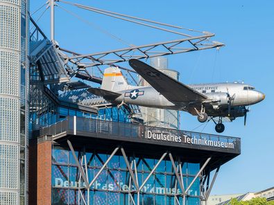 A modern building with a glass front. An airplane is suspended from a steel contruction on the roof.