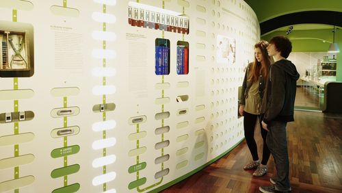 Eine Besucherin und ein Besucher betrachten eine Infografik, die mithilfe von grünen und weißen Lichtfeldern den Weg von der Wirkstoffsuche bis zum fertigen Arzneimittel verdeutlicht.