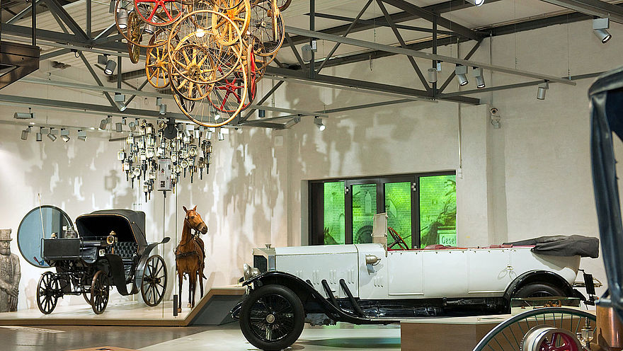 Blick in die Straßenverkehr-Ausstellung: Ein weißes historisches Cabriolet, eine Kutsche mit Pferd und eine Deckeninstallation aus verschiedenen Holz-Rädern.