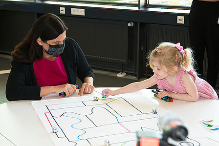 Man sieht eine Frau und ein kleines Mädchen, die an einem Tisch einen kleinen Roboter auf einem Blatt Papier, auf dem eine Linie aufgezeichnet ist, entlangfahren lassen.