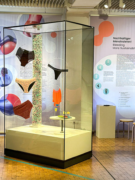 Blick auf die Vitrine im Ausstellungsbereich "Changing Room": Zu sehen sind verschiedene Modelle von Perioden-Unterwäsche, Textil-Binden, Menstruations-Cups und Tampons. Im Hintergrund ist der Schriftzug "Nachhaltiger menstruieren!" zu lesen.