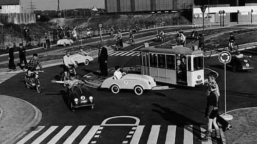 Schwarz-Weiß-Aufnahme, Kinder auf einem Verkehrsparcours in unterschiedlichen Fahrzeugen, im Vordergrund überqueren zwei Kinder einen Zebrastreifen.