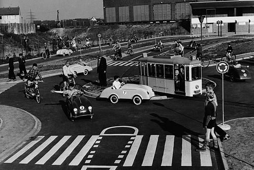 Schwarz-Weiß-Aufnahme, Kinder auf einem Verkehrsparcours in unterschiedlichen Fahrzeugen, im Vordergrund überqueren zwei Kinder einen Zebrastreifen.