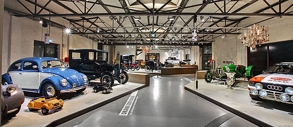 Blick in die Straßenverkehr-Ausstellung: Eine große Halle mit Stahlkonstruktion an der Decke. Ein breiter Gang zwischen niedrigen Podesten mit verschiedenen Fahrzeugen und Objekten. 