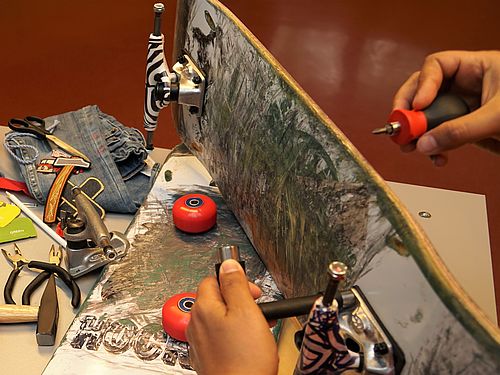 Auf einem Tisch liegen ein kaputtes Skateboard, kaputte Blumentöpfe und eine kaputte Jeans und die Hilfsmittel, um sie zu reparieren.