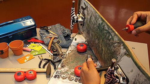 Auf einem Tisch liegen ein kaputtes Skateboard, kaputte Blumentöpfe und eine kaputte Jeans und die Hilfsmittel, um sie zu reparieren.
