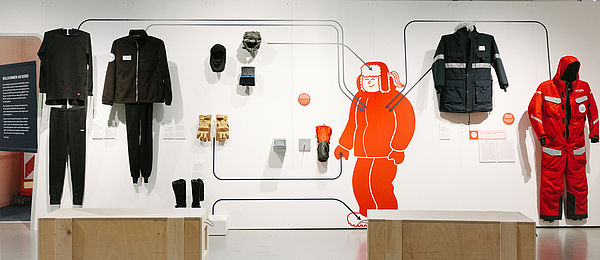 An einer hellen Wand hängen Kleidungsstücke, die man auf einer Polar-Expedition braucht: warme Anoraks, ein roter Ganzkörperanzug, Handschuhe und Mützen.