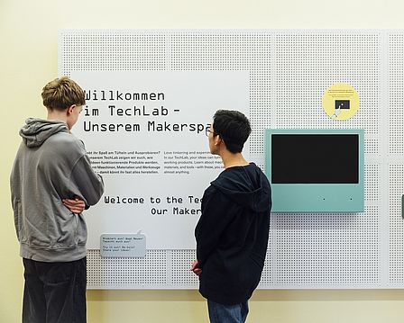 Zwei Jungen stehen vor einer Texttafel auf der die Überschrift „Willkommen im TechLab – unserem Makerspace“ auf Deutsch und Englisch zu erkennen ist. Sie lesen den Text. Daneben hängt ein Monitor mit blauem Rand.