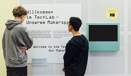 Zwei Jungen stehen vor einer Texttafel auf der die Überschrift „Willkommen im TechLab – unserem Makerspace“ auf Deutsch und Englisch zu erkennen ist. Sie lesen den Text. Daneben hängt ein Monitor mit blauem Rand.