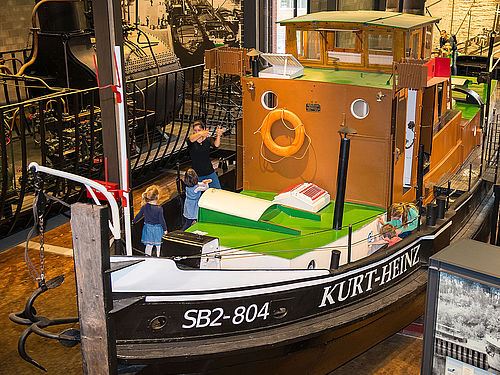 Ein kleines, historisches Dampfschiff steht in der Schifffahrt-Ausstellung. Mehrere Personen stehen auf dem Vorderdeck. Seitlich ist der Schriftzug „SB2 -804“ und „Kurt-Heinz“ zu lesen. 
