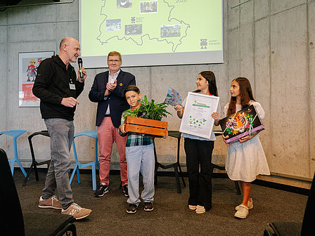 Zwei Männer und drei Kinder stehend vor einer Leinwand. Ein Mann spricht in ein Mikro, ein Kind hält einen Pflanzentopf in der Hand, zwei Kinder eine Urkunde.