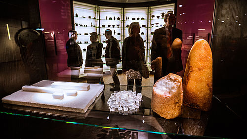 Fünf Personen stehen in einem Raum der Ausstellung „Alles Zucker!“. Im Vordergrund werden in einer Vitrine ein weiß-brauner Zuckerhut, Teile von Rohrzucker, Kandis-Zuckerkristalle und gepresster Zucker gezeigt. Im Hintergrund befinden sich deckenhohe Vitrinen mit Zuckerdosen.
