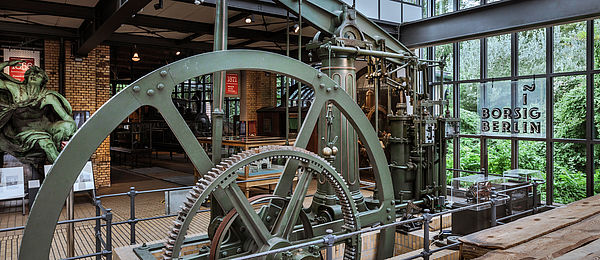 Blick auf das Rad einer grünen Dampfmaschine. Es ist teilweise im Boden versenkt und wird durch eine eiserne Absperrung gesichert. Durch das verglaste Dach und ein bodentiefes Fenster fällt Licht in den Ausstellungsraum. Auf einem Leuchtschild vor der Fensterfront steht „Borsig Berlin“.