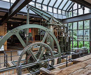 Blick auf das Rad einer grünen Dampfmaschine. Es ist teilweise im Boden versenkt und wird durch eine eiserne Absperrung gesichert. Durch das verglaste Dach und ein bodentiefes Fenster fällt Licht in den Ausstellungsraum. Auf einem Leuchtschild vor der Fensterfront steht „Borsig Berlin“.