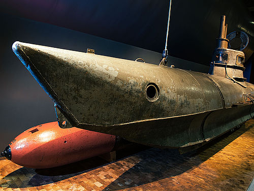Schrägansicht des Einmann-U-Boots Biber. Das U-Boot ist etwa neun Meter lang, verfügt über einen kleinen Turm mit Aussichtsfenstern und besteht aus grauem Metall. Die Nase des U-Boots ist nach oben gebogen. Dahinter liegt ein roter Torpedo.