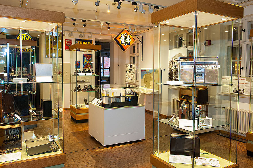 Eine Ansicht der Ausstellung Nachrichtentechnik. Mehrere Glas-Vitrinen zeigen Radios, in der Mitte steht ein Modell des Vox Hauses. 