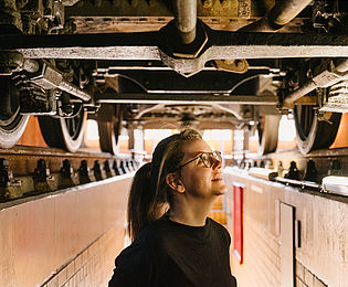 Eine junge Frau steht in einem schmalen Gang unter einer historischen Lokomotive. Der Boden der Lok mit Rädern und Fahrwerk ist über ihr zu erkennen.