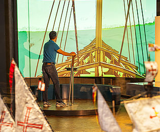 Ein Mann steht auf einem Podest und steuert ein Segelschiff, das vor ihm auf die Wand projiziert wird.