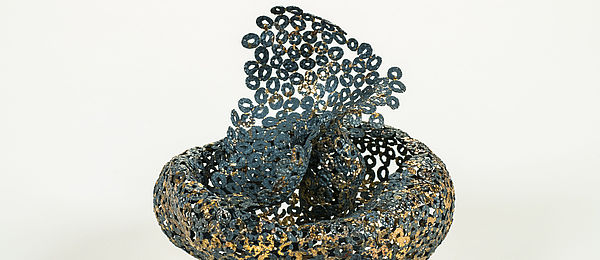 Schwarze Brosche mit Goldflecken aus einer Vielzahl kleiner Ösen. Die Ösen wurden wie beim 3D-Druck von innen nach außen zu einem voluminösen Körper aufgebaut.