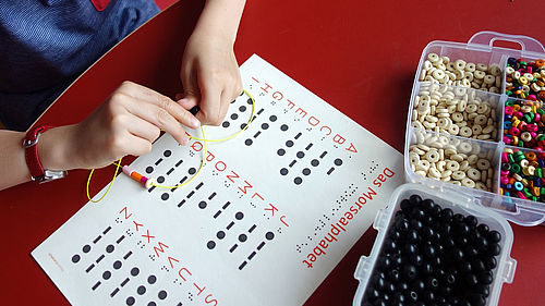 Ein Mädchen sitzt an einem Tisch und fädelt bunte Holzperlen auf einen gelben Faden. Auf dem Tisch liegt ein Blatt, auf dem das Morsealphabet erklärt wird. Daneben befinden sich zwei durchsichtige Behältnisse mit weißen, schwarzen und bunten Perlen. Von dem Mädchen sind nur die Hände zu sehen.