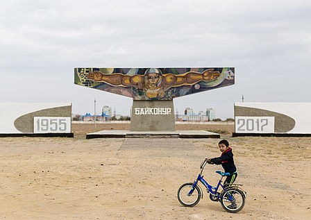 Ein kleiner Junge schiebt sein Fahrrad über einen großen sandigen Platz. Im Hintergrund steht ein großes Denkmal, darauf der Kopf einen Kosmonauten, der seine Arme weit ausbreitet.