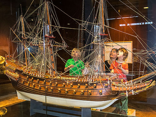 Eine Frau und drei Kinder stehen vor einer Vitrine mit einem etwa drei Meter breiten und zwei Meter hohen Modell eines dreimastigen Segelschiffs. Das Modell besteht aus Holz, der Bauch des Schiffs ist weiß lackiert.