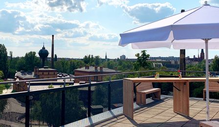 Auf der Terrasse, die zur Eventfläche AUSGUCK gehört, stehen leichte sommerliche Holzmöbel und ein weißer Landhausschirm. Man hat einen weiten Blick über das Museumsgelände, Richtung Lokschuppen, bis zum Wasserturm.