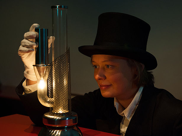 Eine Frau in schwarzem Anzug, mit weißen Handschuhen und mit schwarzem Zylinder experimentiert in einem abgedunkelten Raum. Auf der roten Tischplatte vor ihr steht ein Glasrohr, aus dem ein weiteres Glasrohr abzweigt wie ein Arm eines Kaktus. Auf diesem Rohr sitzt ein silbernes metallisches Rohr, darunter brennt eine kleine Flamme, etwa so groß wie von einer Kerze. Die Frau hält das silberne Rohr in einer Hand und schaut gespannt auf die Flamme.