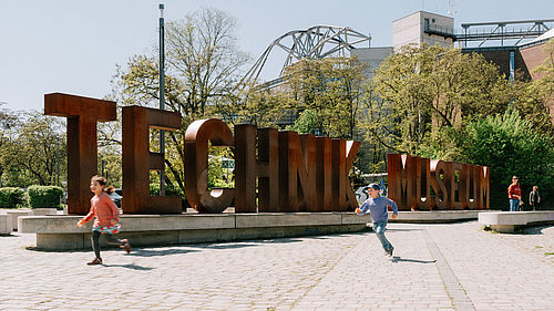 Zwei Kinder rennen über den Vorplatz des Deutschen Technikmuseums mit der Wortskulptur "Technik Musem". 