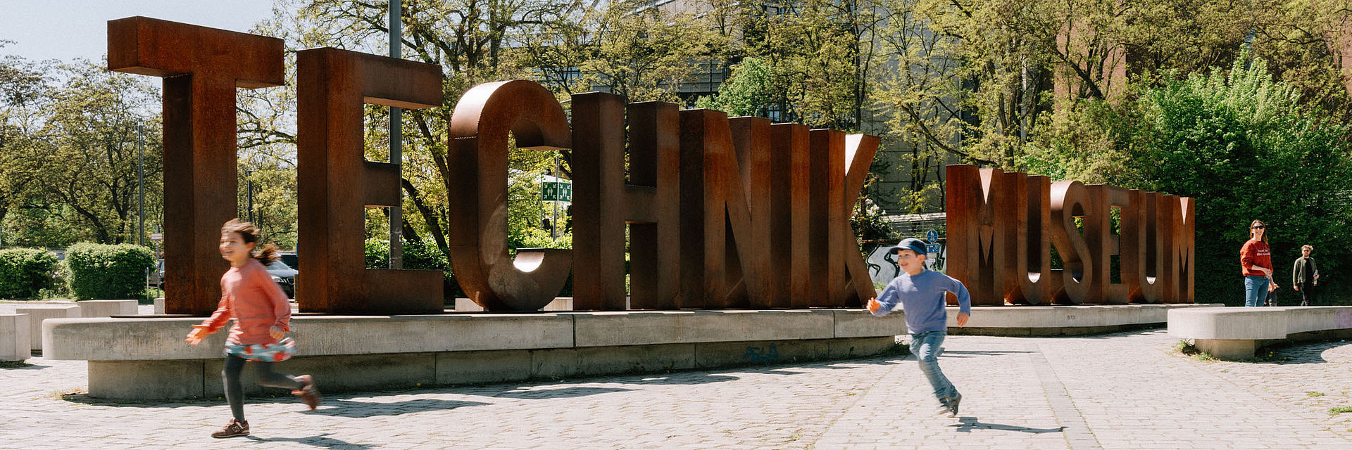 Zwei Kinder rennen über den Vorplatz des Deutschen Technikmuseums mit der Wortskulptur "Technik Musem". 