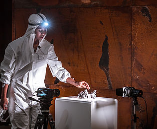 Eine Frau mit dunkler Hautfarbe, in einen weißen Schutzanzug mit Stirnlampe gekleidet, berührt mit einer Hand einen Haufen Staub, der auf einem Sockel liegt. Im Hintergrund ist eine braune Metallwand zu sehen.