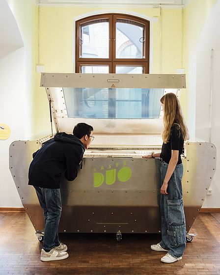 Ein Mädchen und ein Junge betrachten ein großes, silberfarbenes technisches Gerät. Auf dem gerät steht „LaserDuo“. Der Junge beugte sich über die Öffnung des Geräts, das Mädchen steht neben ihm und betrachtet das Innere des Geräts.
